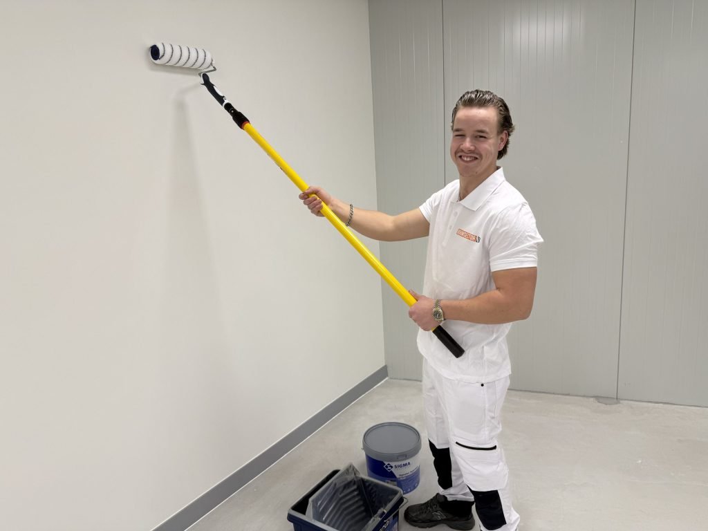 Lachende schilder in witte werkkleding saust een lichte muur, met een emmer Sigma-verf en een verfbak op de vloer