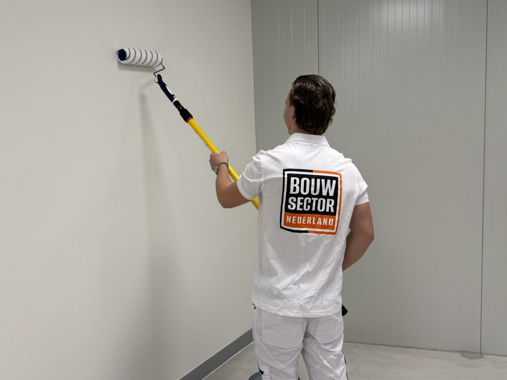 Schilder van Bouwsector Nederland rolt met een gele telescoopsteel een kale witte wand in een lege ruimte