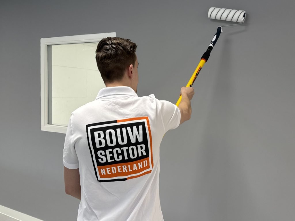 Schilder met het Bouwsector Nederland-logo op zijn rug zet de roller hoog op een grijze wand naast een raampje