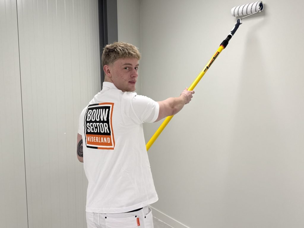 Schilder kijkt achterom terwijl hij met een roller op een lange steel een lichte wand in een gang sauzt