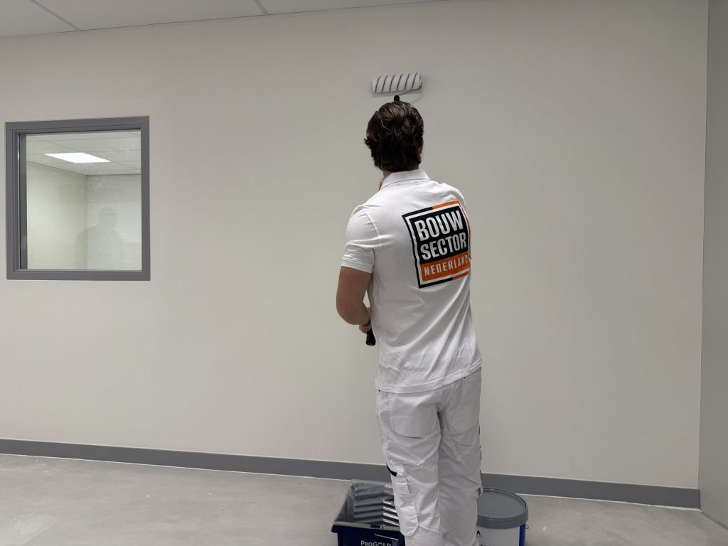 Schilder in wit werkpak rolt een lichte wand tot vlak onder het plafond, met verfbak en emmer aan zijn voeten