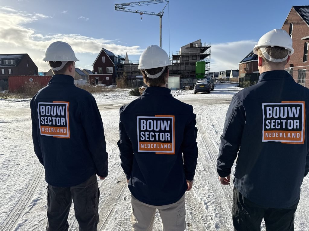Drie vakmensen van Bouwsector Nederland met bouwhelm staan op een nieuwbouwlocatie met een kraan op de achtergrond